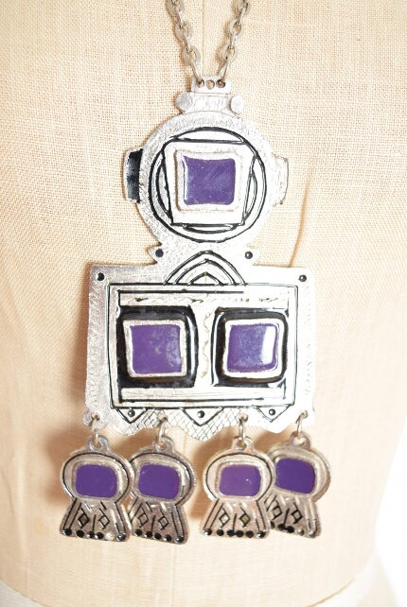 May include: A silver necklace with a large pendant featuring a geometric design with purple accents. The pendant has a square shape with two smaller squares inside, and the necklace has five smaller purple squares hanging from the bottom.