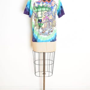 May include: A white t-shirt with a colorful tie-dye pattern and a graphic illustration on the back. The illustration depicts a skeleton character looking through a telescope, surrounded by birds and a window.