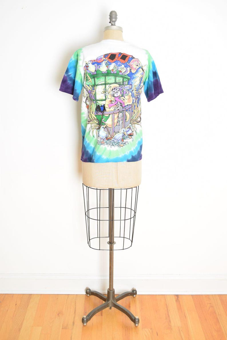 May include: A white t-shirt with a colorful tie-dye pattern and a graphic illustration on the back. The illustration depicts a skeleton character looking through a telescope, surrounded by birds and a window.