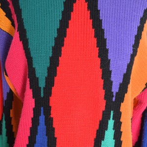 May include: A colorful sweater with a geometric pattern. The sweater features a black crew neck and black diamond shapes outlined in black. The diamonds are filled with red, pink, green, purple, and orange. The sleeves are visible.