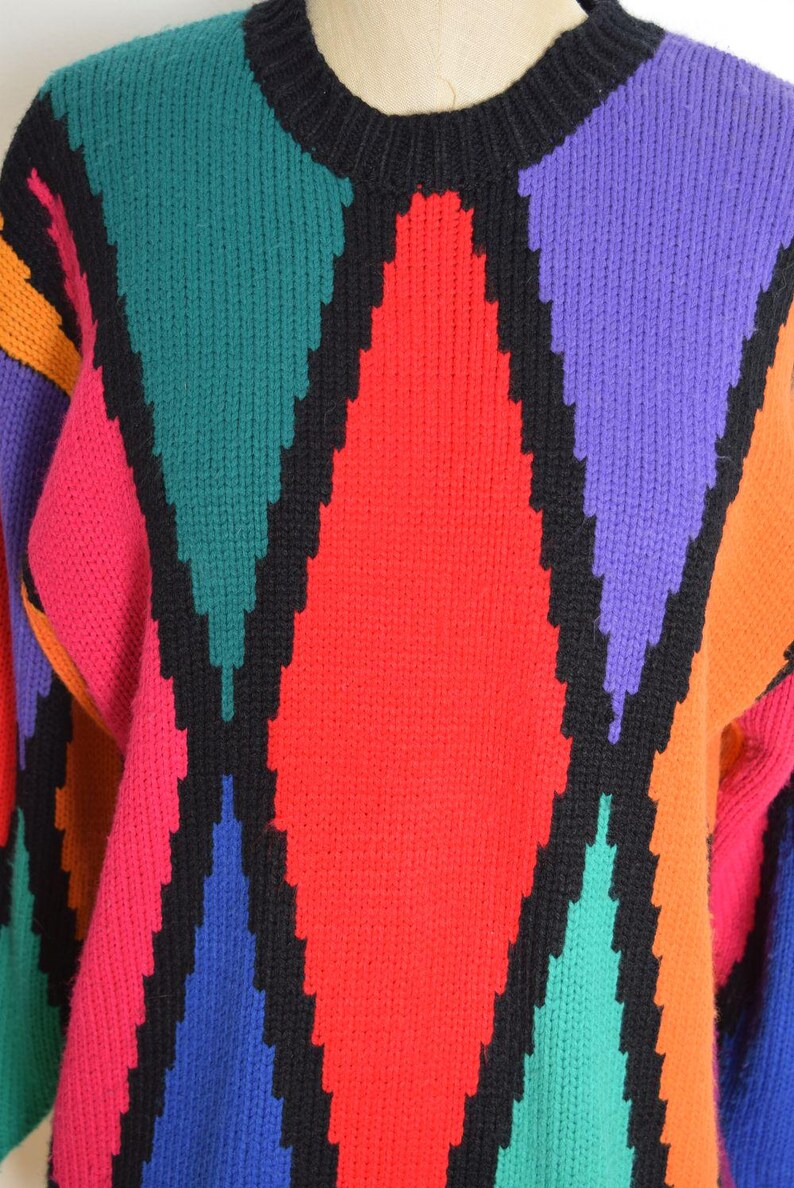 May include: A colorful sweater with a geometric pattern. The sweater features a black crew neck and black diamond shapes outlined in black. The diamonds are filled with red, pink, green, purple, and orange. The sleeves are visible.