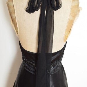 May include: Black halter-neck dress with a flowing skirt, displayed on a mannequin. The dress features a beaded waist detail and a sheer, flowing scarf that ties at the neck. The fabric has a satin sheen.