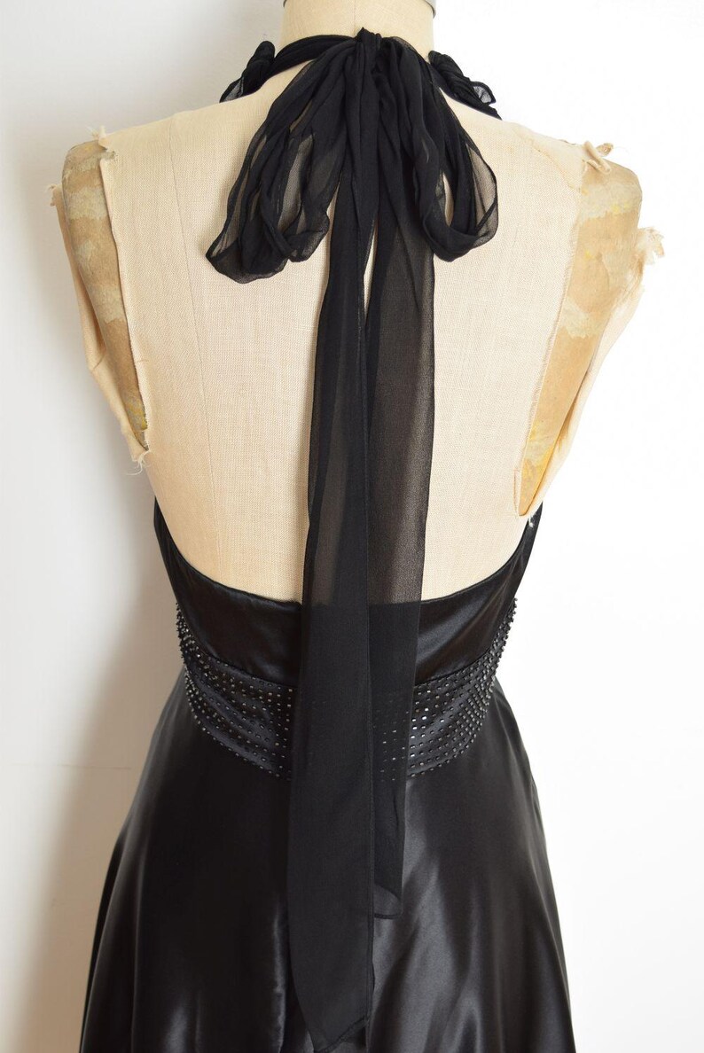 May include: Black halter-neck dress with a flowing skirt, displayed on a mannequin. The dress features a beaded waist detail and a sheer, flowing scarf that ties at the neck. The fabric has a satin sheen.
