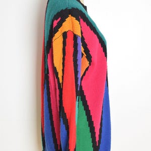 May include: A colorful, vintage sweater with a geometric pattern. The sweater features a black collar and trim, with bold blocks of teal, orange, red, pink, purple, green, and blue. The sweater is displayed on a mannequin.