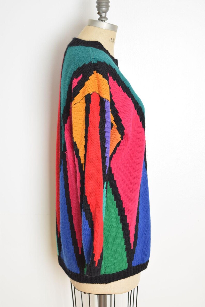 May include: A colorful, vintage sweater with a geometric pattern. The sweater features a black collar and trim, with bold blocks of teal, orange, red, pink, purple, green, and blue. The sweater is displayed on a mannequin.