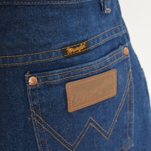 May include: Close-up of the back pocket of blue denim jeans. The pocket features a brown leather patch with the Wrangler logo and copper rivets. The Wrangler brand name is also visible on a small tag above the pocket.