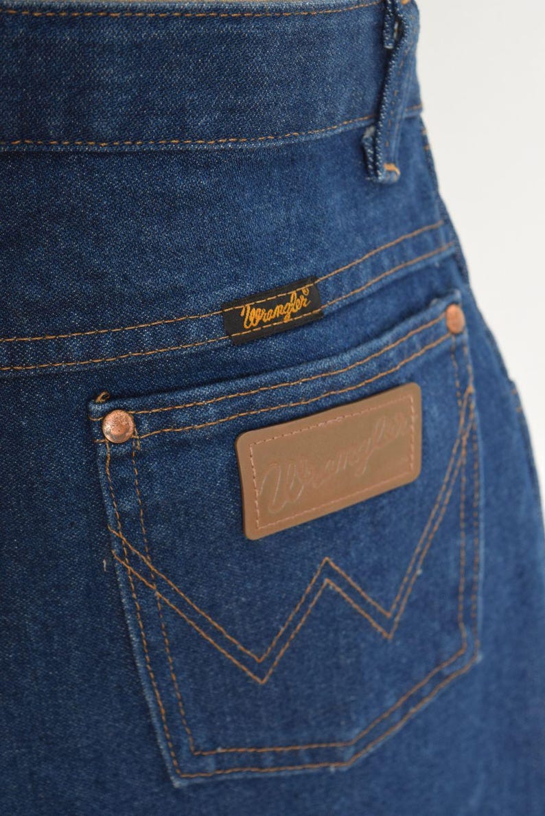 May include: Close-up of the back pocket of blue denim jeans. The pocket features a brown leather patch with the Wrangler logo and copper rivets. The Wrangler brand name is also visible on a small tag above the pocket.