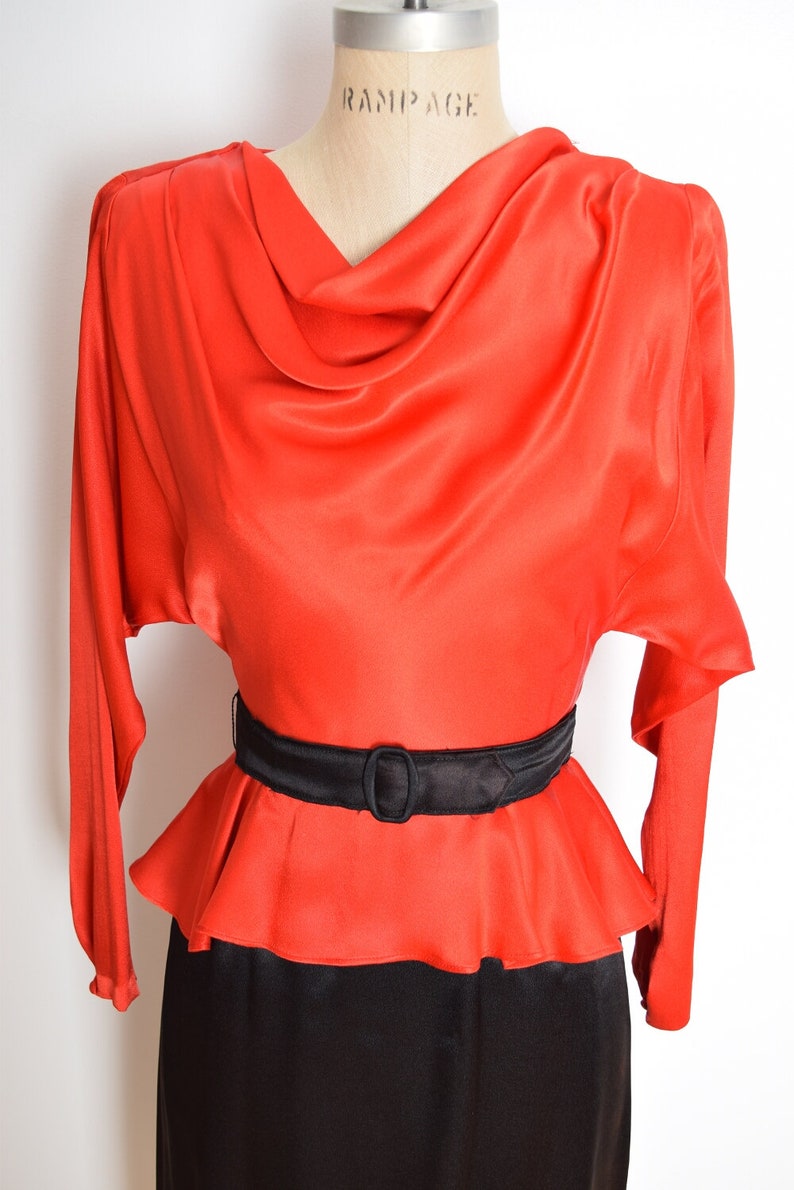 May include: A red satin dress with a cowl neck and a black belt. The dress has a fitted bodice and a flowing skirt.
