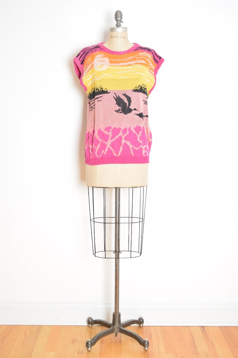 May include: A pink, yellow, and orange sleeveless sweater with a black and white graphic print of a sunset and birds. The sweater is on a mannequin.
