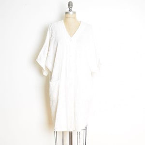 May include: A white, textured, knee-length robe with a V-neckline, short wide sleeves, and a front button closure. The garment features a vertical ribbed pattern throughout and includes two large patch pockets on the front lower section.