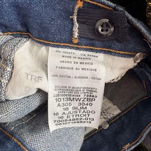 May include: Close-up of blue denim jeans with a button and adjustable waistband. A white tag displays text including "MADE IN MEXICO", "100% COTTON", and size information. The jeans have orange stitching.