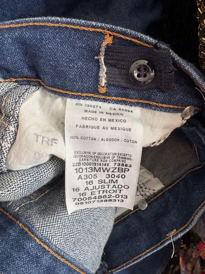 May include: Close-up of blue denim jeans with a button and adjustable waistband. A white tag displays text including "MADE IN MEXICO", "100% COTTON", and size information. The jeans have orange stitching.