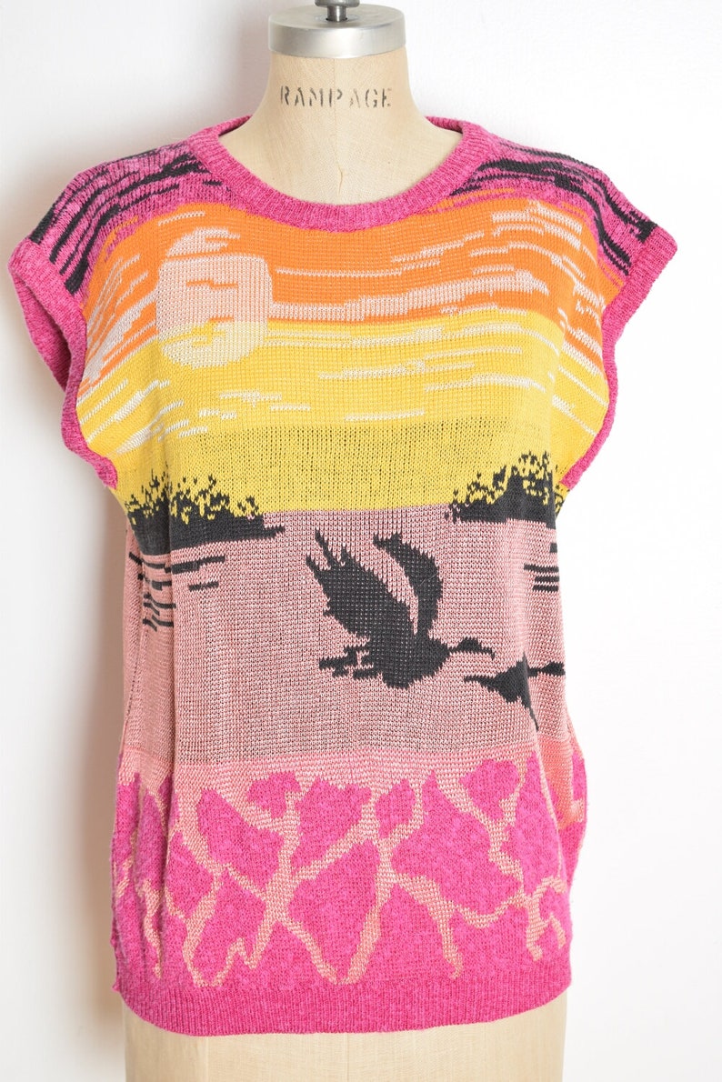 May include: A pink, yellow, orange, and black sleeveless knitted sweater with a sunset and bird design. The sweater has the word "Rampage" on the label.