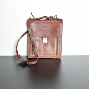 May include: A brown leather satchel with a flap closure and a gold-colored clasp. The bag has a shoulder strap and a top handle. The satchel is rectangular and appears to be vintage.