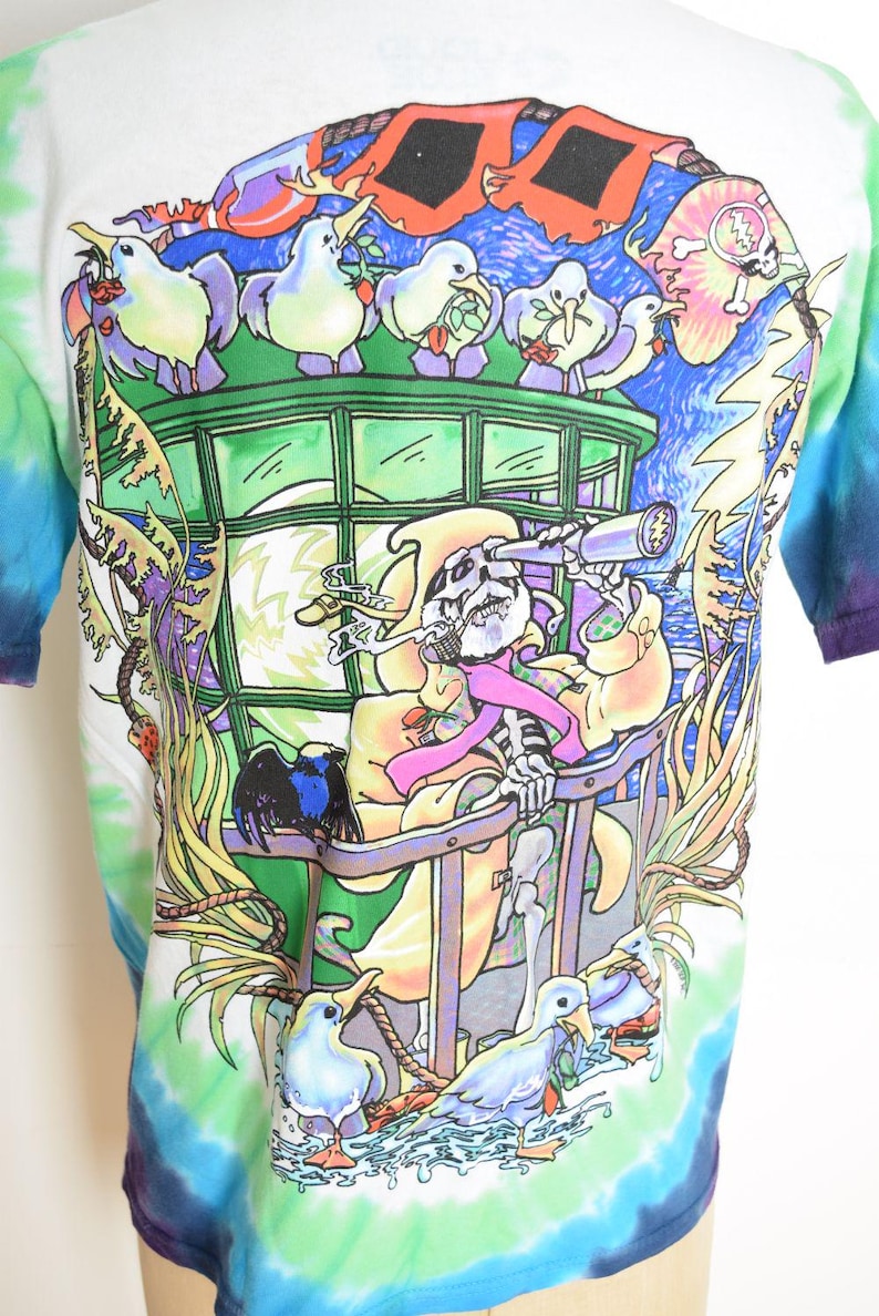 May include: Tie-dye t-shirt with a colorful graphic. The design features a skeleton with a telescope, surrounded by cartoon birds and a vibrant, whimsical landscape. The shirt has a white base with blue, green, and purple tie-dye.