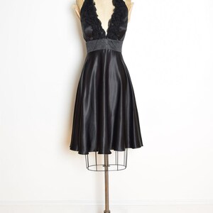 May include: A black satin halter dress displayed on a vintage dress form. The dress features a V-neckline with a ruffled trim and a beaded waist. The skirt flares out from the waist, creating a classic silhouette.