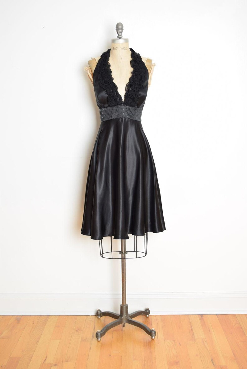May include: A black satin halter dress displayed on a vintage dress form. The dress features a V-neckline with a ruffled trim and a beaded waist. The skirt flares out from the waist, creating a classic silhouette.