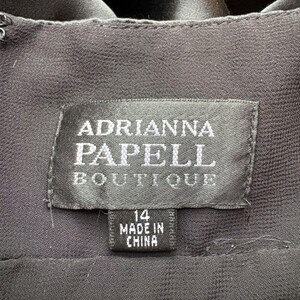 May include: Close-up of a black garment label. The label reads "ADRIANNA PAPELL BOUTIQUE" in silver lettering. Below the main label is a smaller tag indicating size 14 and "MADE IN CHINA". The fabric appears to be a dark, flowing material.