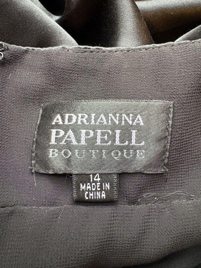 May include: Close-up of a black garment label. The label reads "ADRIANNA PAPELL BOUTIQUE" in silver lettering. Below the main label is a smaller tag indicating size 14 and "MADE IN CHINA". The fabric appears to be a dark, flowing material.
