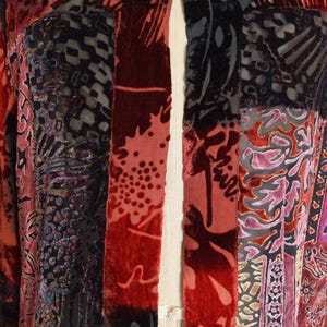 May include: A patchwork jacket with a variety of patterned velvet panels in shades of red, black, and purple. The jacket is open in the front and has a unique, artistic design. The garment is displayed on a mannequin.