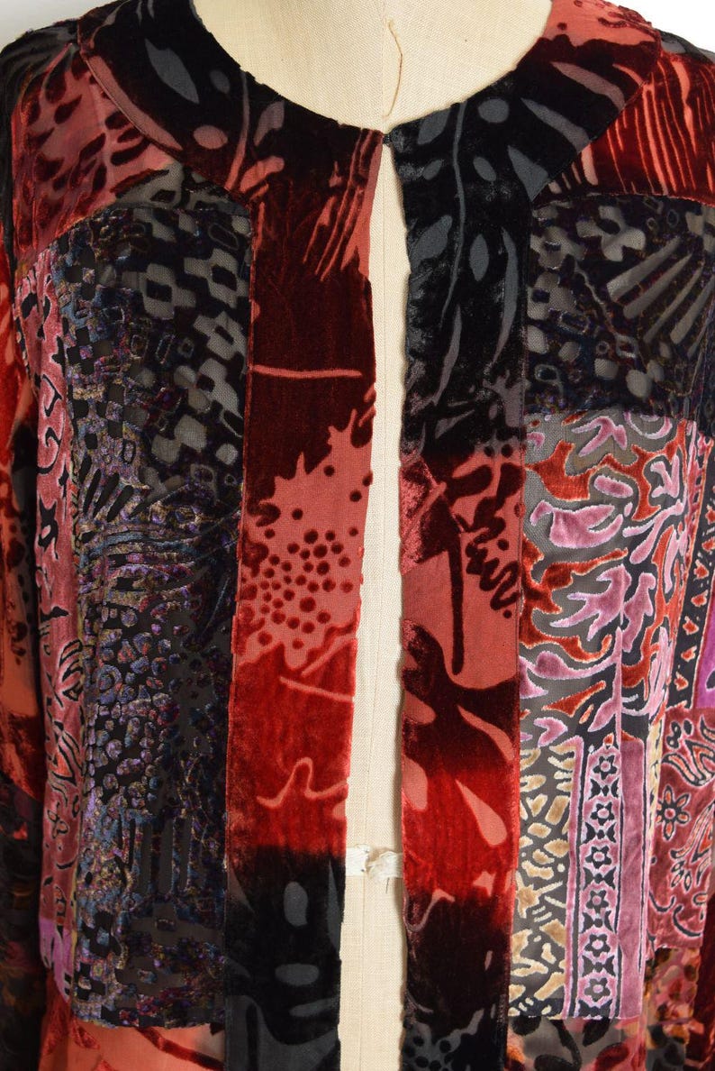 May include: A patchwork jacket with a variety of patterned velvet panels in shades of red, black, and purple. The jacket is open in the front and has a unique, artistic design. The garment is displayed on a mannequin.