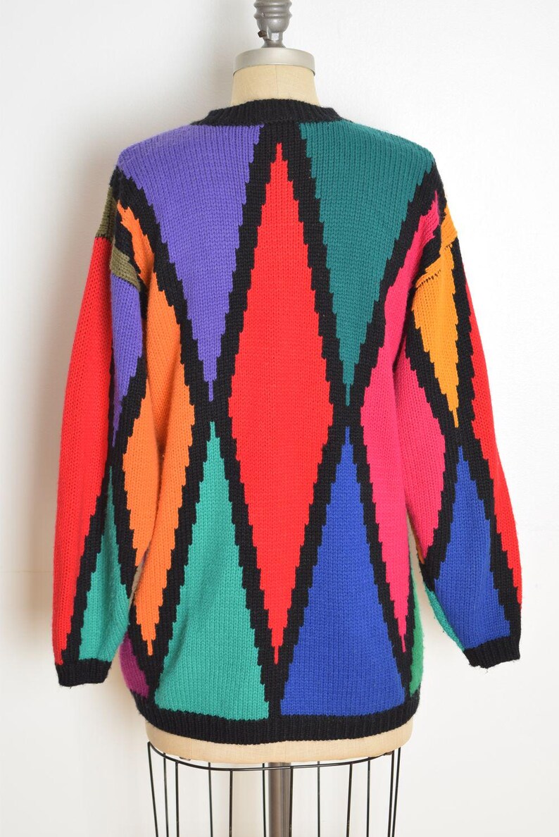 May include: A colorful, vintage sweater with a geometric diamond pattern. The sweater features a black neckline and trim, and a vibrant array of colors including red, purple, green, orange, and blue. The sweater is displayed on a mannequin.
