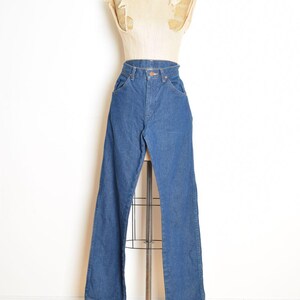 May include: Blue denim jeans with a high waist and flared leg, displayed on a vintage dress form. The jeans have classic five-pocket styling and a button and zipper closure. The mannequin is on a metal stand.