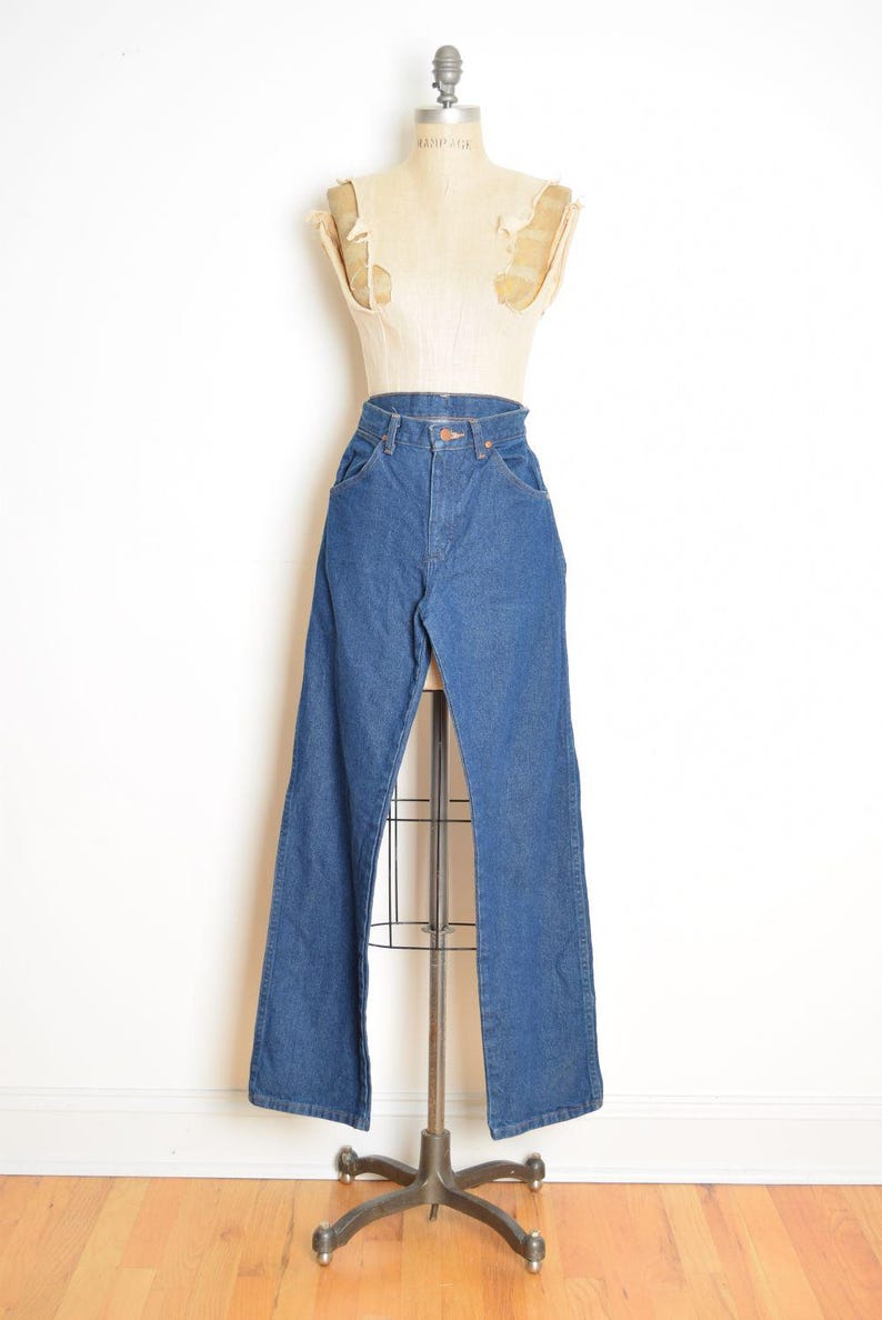 May include: Blue denim jeans with a high waist and flared leg, displayed on a vintage dress form. The jeans have classic five-pocket styling and a button and zipper closure. The mannequin is on a metal stand.