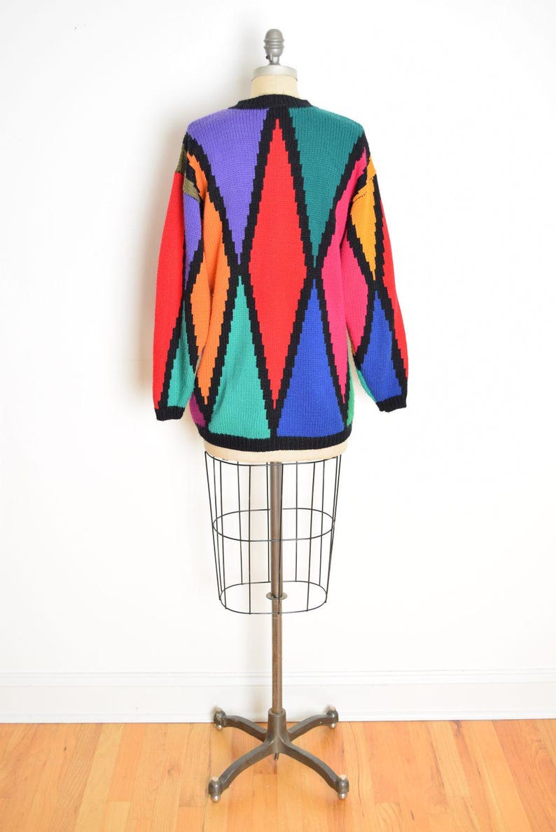 May include: A colorful sweater with a geometric diamond pattern in red, purple, green, blue, orange, and pink, outlined in black. The sweater is displayed on a wire dress form against a white background.