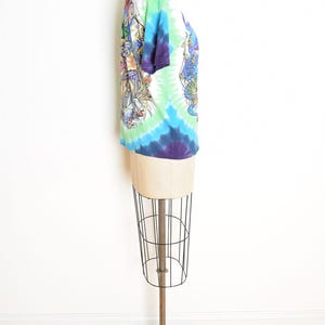 May include: A tie-dye t-shirt with a white collar and sleeves, displayed on a mannequin. The shirt features a vibrant swirl of colors, including blue, green, and purple. Detailed graphic designs are visible on the front and back of the shirt.