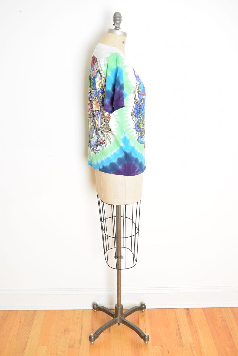 May include: A tie-dye t-shirt with a white collar and sleeves, displayed on a mannequin. The shirt features a vibrant swirl of colors, including blue, green, and purple. Detailed graphic designs are visible on the front and back of the shirt.