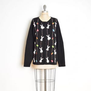 May include: Black cardigan sweater with white bunny and colorful egg designs. The sweater has a round neckline and button-up front. White fluffy accents and beaded strings add detail to the Easter-themed knitwear.