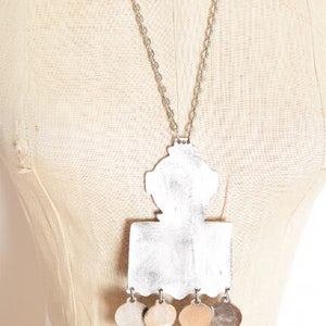 May include: A silver necklace with a large, ornate pendant featuring a geometric design and five smaller, round pendants hanging below. The pendant is silver and has a textured surface.