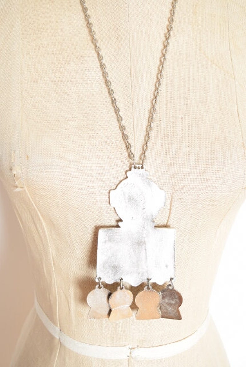 May include: A silver necklace with a large, ornate pendant featuring a geometric design and five smaller, round pendants hanging below. The pendant is silver and has a textured surface.