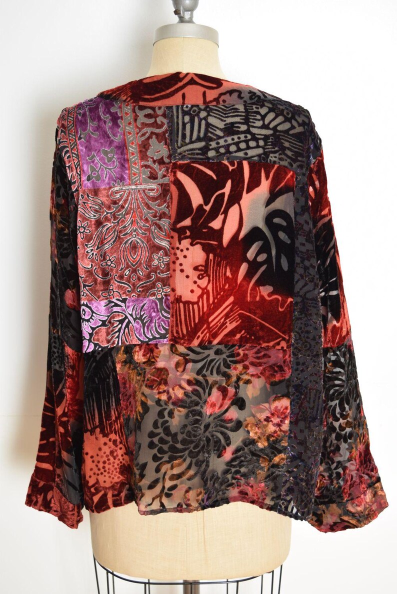 May include: A patchwork velvet top in shades of red, purple, and black. The long-sleeved garment features a variety of patterned fabrics, including floral and geometric designs. The top is displayed on a mannequin.