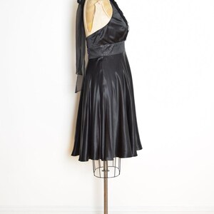 May include: A black satin dress displayed on a dress form. The dress features a halter neckline with a ruffled trim, a sequined belt, and a flared skirt. A sheer black scarf drapes over the shoulder.
