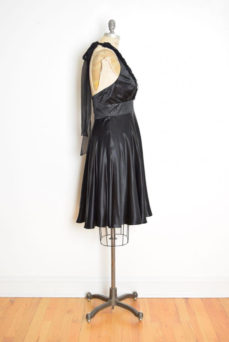 May include: A black satin dress displayed on a dress form. The dress features a halter neckline with a ruffled trim, a sequined belt, and a flared skirt. A sheer black scarf drapes over the shoulder.
