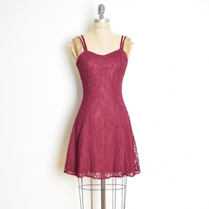 Peut inclure: Une robe sans manches arrivant au genou de couleur bordeaux profond, dotée d'un motif floral en dentelle superposé sur toute la surface. Le vêtement présente une encolure en cœur, de fines bretelles et un corsage ajusté s'évasant en jupe.