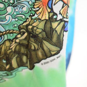 May include: Close-up of a tie-dye shirt with a colorful graphic design. The design features a detailed illustration of a mythical creature with orange and yellow wings, set against a backdrop of water and rocks. The text "&copy; 2000 GDP, INC" is visible.