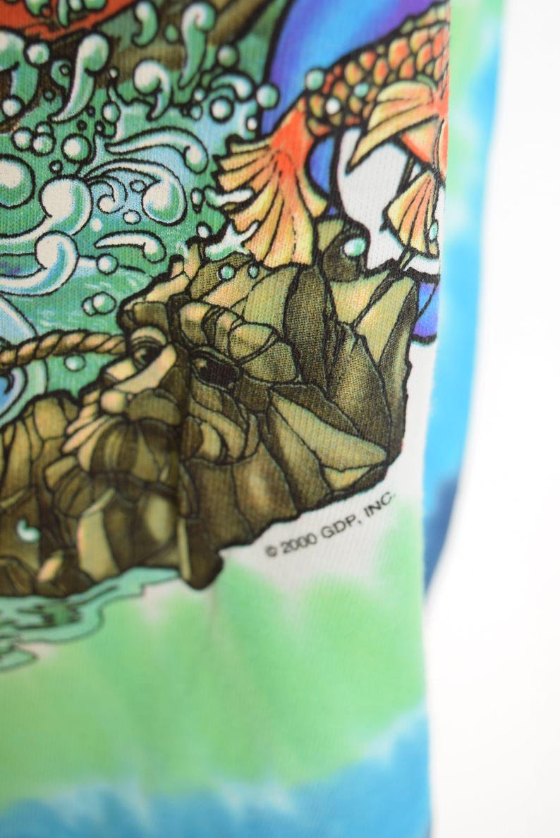 May include: Close-up of a tie-dye shirt with a colorful graphic design. The design features a detailed illustration of a mythical creature with orange and yellow wings, set against a backdrop of water and rocks. The text "&copy; 2000 GDP, INC" is visible.