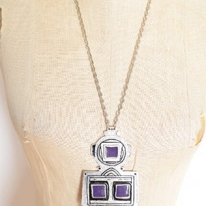 May include: A silver necklace with a large pendant featuring a geometric design with purple glass accents. The pendant has a square shape with two smaller squares in the center and three dangling charms.