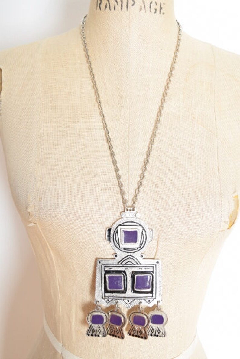 May include: A silver necklace with a large pendant featuring a geometric design with purple glass accents. The pendant has a square shape with two smaller squares in the center and three dangling charms.