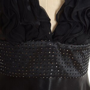 May include: A black evening dress with a V-neckline and a beaded waist. The bodice features ruffled fabric, and the skirt is made of a smooth, shiny material. The dress is displayed on a mannequin.