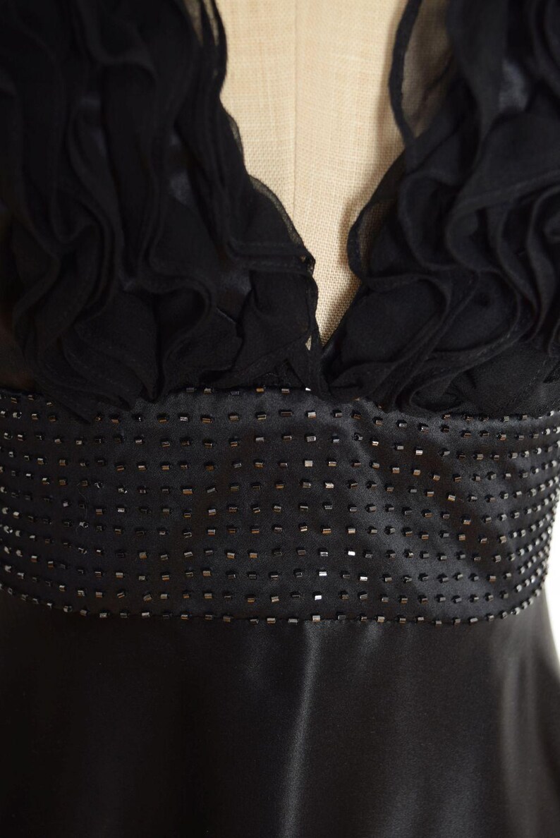 May include: A black evening dress with a V-neckline and a beaded waist. The bodice features ruffled fabric, and the skirt is made of a smooth, shiny material. The dress is displayed on a mannequin.