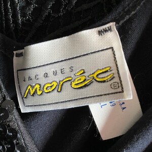 May include: A white tag with black text that reads "Jacques Morel" in a yellow script font. The tag is attached to a black garment with sequins.