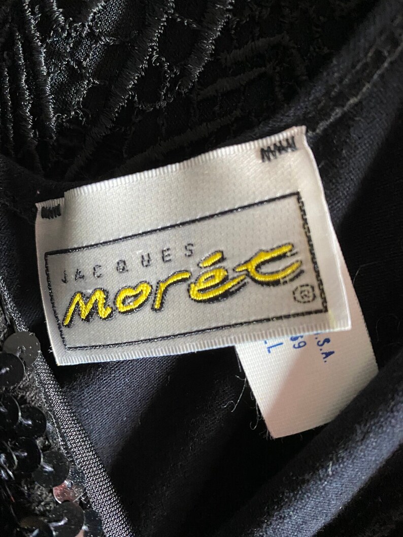 May include: A white tag with black text that reads "Jacques Morel" in a yellow script font. The tag is attached to a black garment with sequins.