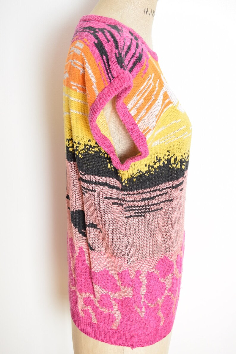 May include: A pink, orange, yellow, black and white striped knit sweater with a short sleeve design. The sweater has a floral pattern at the bottom.