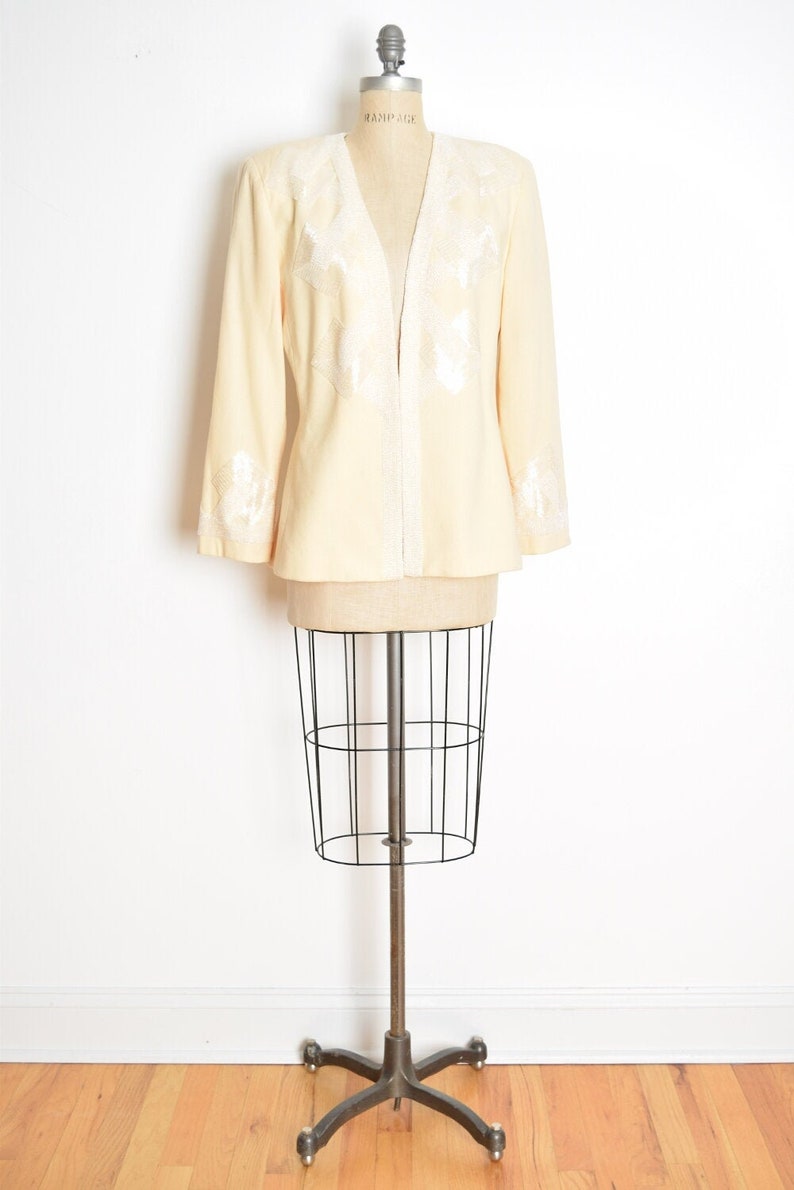May include: A cream-colored blazer with white beaded embellishments on the front and sleeves. The blazer is on a dress form.