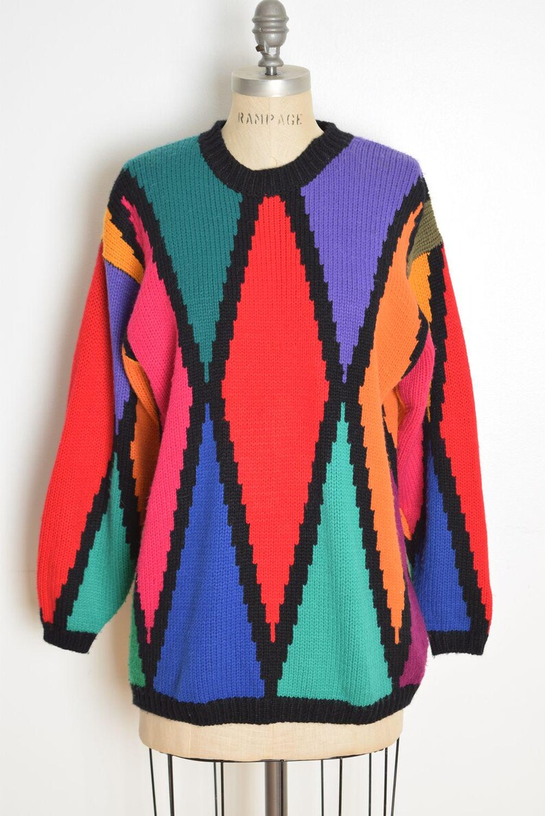 May include: A vintage, oversized sweater with a bold geometric pattern. The sweater features large diamond shapes in red, purple, green, blue, orange, and pink, outlined in black. The neckline and cuffs are black. The brand name "RAMPAGE" is visible on the mannequin.
