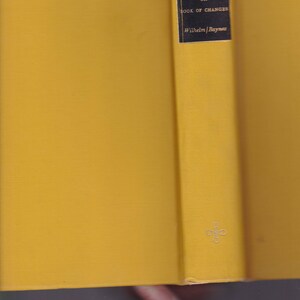 May include: Open book with yellow covers. The spine features a black label with white text: "THE I CHING OR BOOK OF CHANGES" and "Wilhelm/Baynes". A small, silver emblem is visible near the bottom of the spine.
